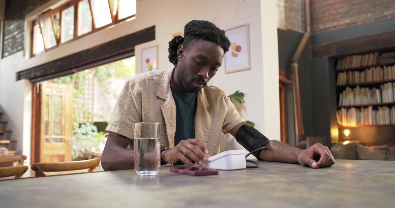 African man in shirt fitting cuff, pressing monitor, checking blood pressure at kitchen with phone
