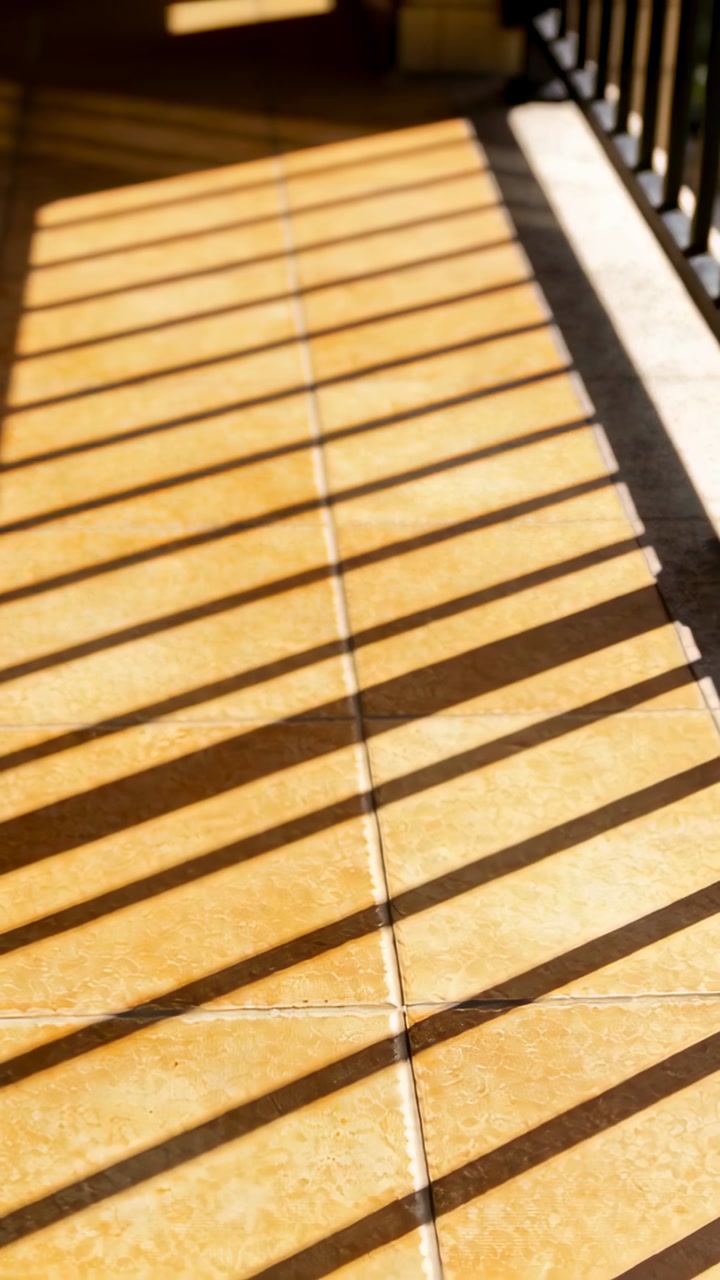 Vertical video: Sun casting angled shadow bars from metal rail on balcony tiles and sunlit ledge