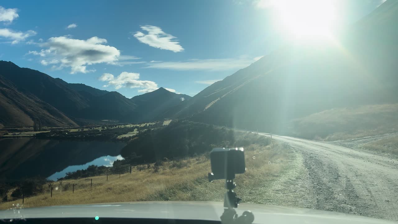Windshield wiper sweeps across car window, revealing sunlit mountain lake road in Queenstown, New Zealand