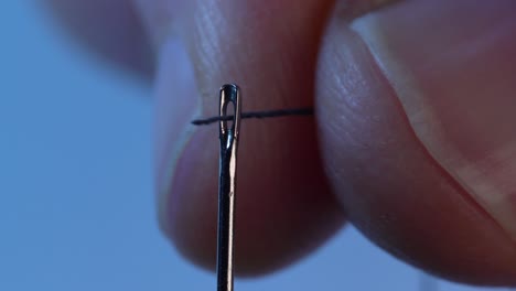 thread hold between index finger and thumb being smoothly guided through the eye of the needle against solid blue background