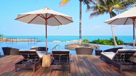 Amazing-Exotic-Infinity-Pool-Lounge-with-Coconut-Trees-on-the-Rocky-Island-in-Bali,-Deck-chairs,-and-white-Umbrellas-with-small-tables-in-front