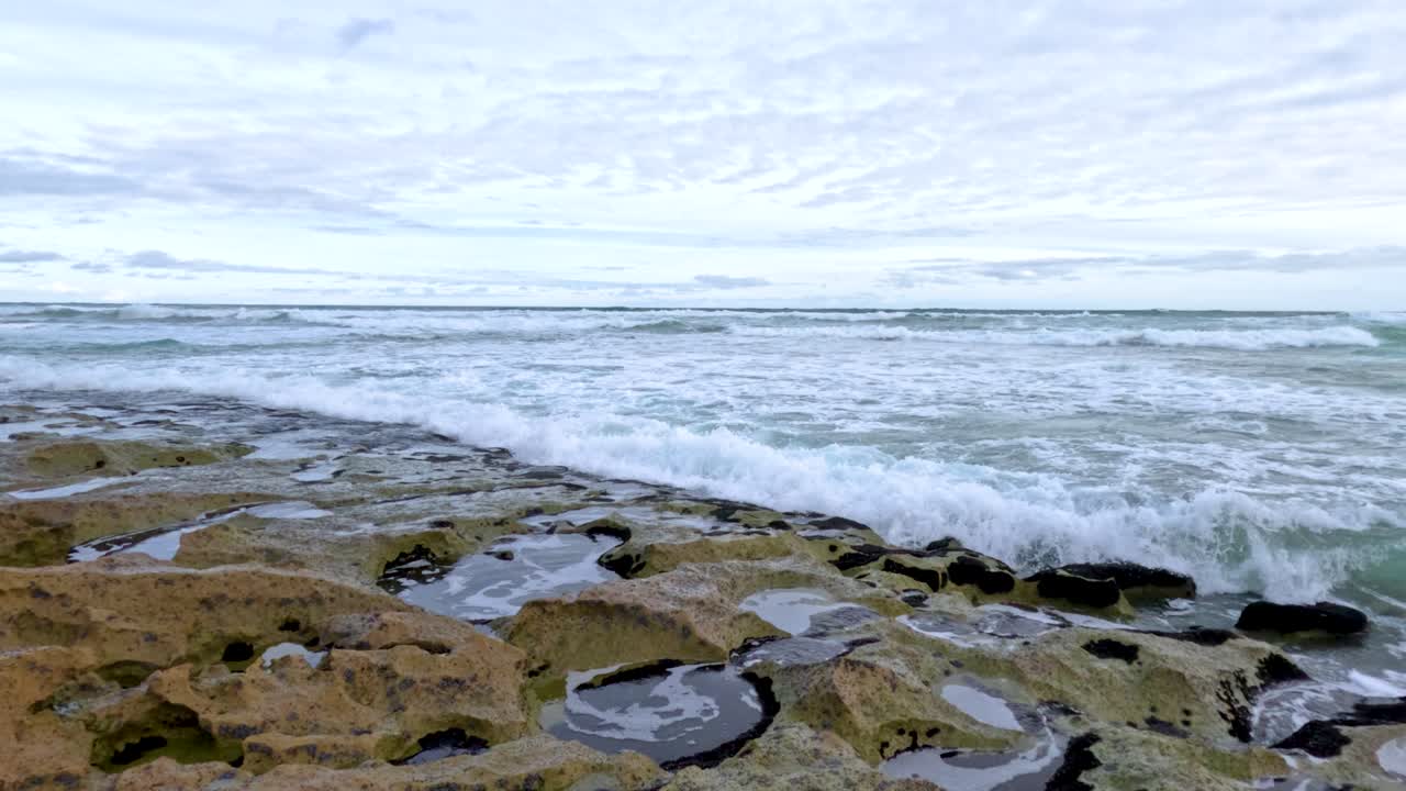 Dynamic ocean waves crash against rocky formations under a cloudy sky, showcasing nature's power and beauty