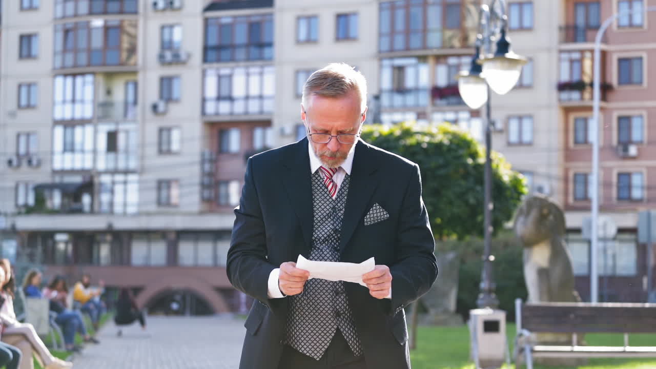 Mature businessman with a sheet of paper on the city background. Portrait of an old man in elegant costume reading paper and becomes very happy outdoors.