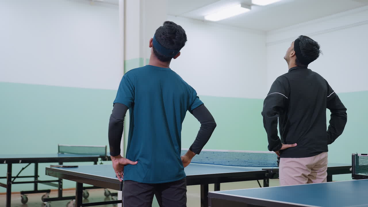Back view of two friends in sportswear performing neck flexibility exercise near tennis table, warming up muscles and joints to prepare for sports activity inside brightly lit indoor hall