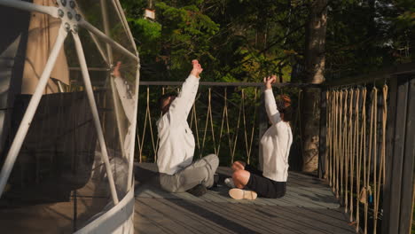 couple does yoga exercise at glamping. instructor shows hand raising asana to student sitting on eco hotel terrace. people practice mindfulness together