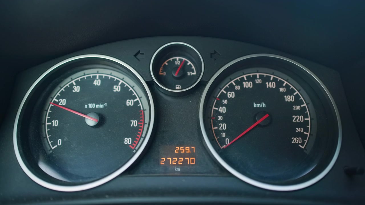 Close view of car instrument cluster inside dark interior, showing two large circular dials, central fuel gauge, digital odometer, and red needles gently shifting across marked scales with clear