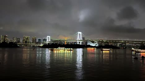 Beleuchtete-Stadtlandschaft-Mit-Einer-Beleuchteten-Brücke-Und-Spiegelungen-Auf-Dem-Wasser-Bei-Nacht