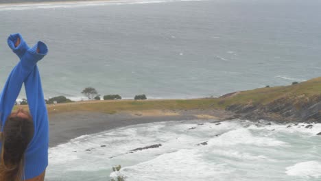 Vista-Trasera-De-Una-Mujer-Con-Sudadera-Azul-Recortada-Levantando-Las-Manos-Y-Admirando-La-Pequeña-Playa-De-Arena-Y-Guijarros-Desde-El-Mirador-De-Crescent-Head