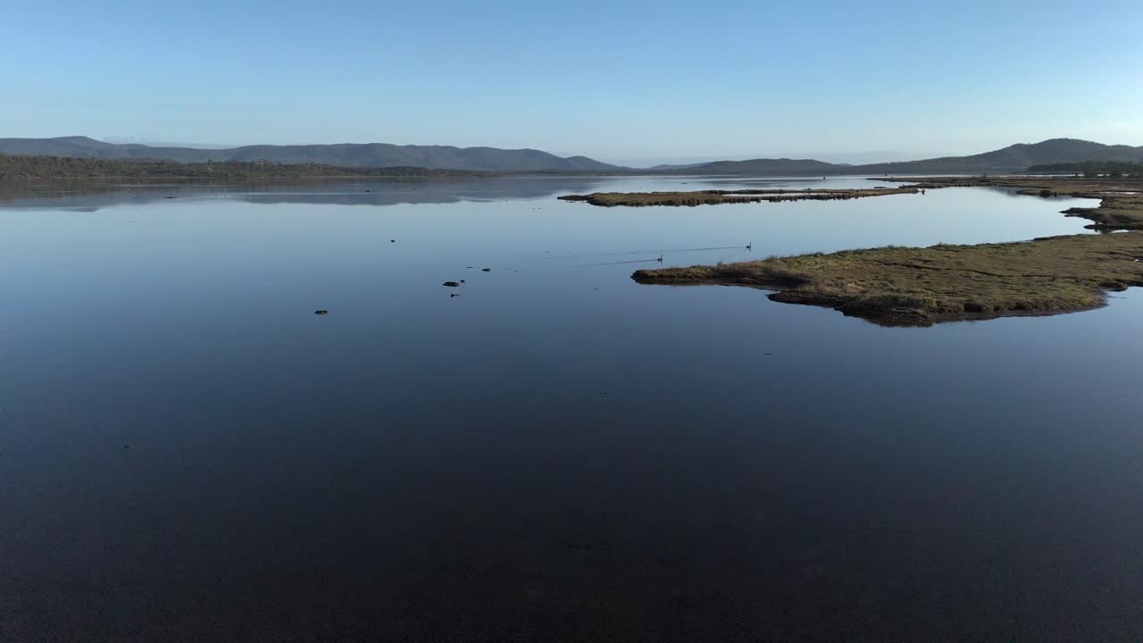 Aerial flyover Tasmanian reserve lake area with islands and mountains during sunny day and blue sky