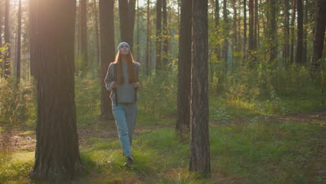 young woman walking through a tranquil forest, holding backpack straps as she gazes thoughtfully to the left, evening sunlight casts a warm glow, tall trees and lush greenery line her path