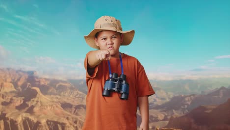 asian boy with a hat and binoculars pointing his finger towards you and furiously speaking against blaming you while traveling at the top of mountain. boy researcher, travel tourism adventure concept