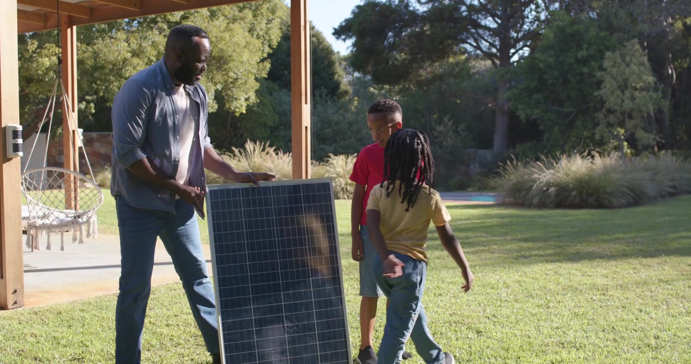 Afroamerikansk far med to drenge bærer solpanel ud på solrig græsplæne, placerer og forklarer.