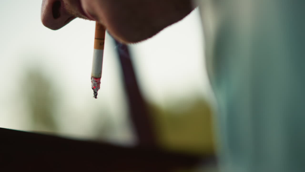 primo piano di un uomo caucasico, sigaretta, cenere che cade, dettaglio intenso della punta con braci incandescenti, cenere e residui sospesi, profondità di campo ridotta con vegetazione sfocata, texture cinematografica suggestiva e quieta tensione