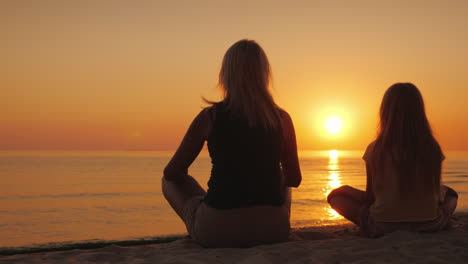 A-Woman-With-Her-Daughter-Sitting-Side-By-Side-On-The-Sand-In-A-Lotus-Pose-Looking-At-The-Sunset-Ove