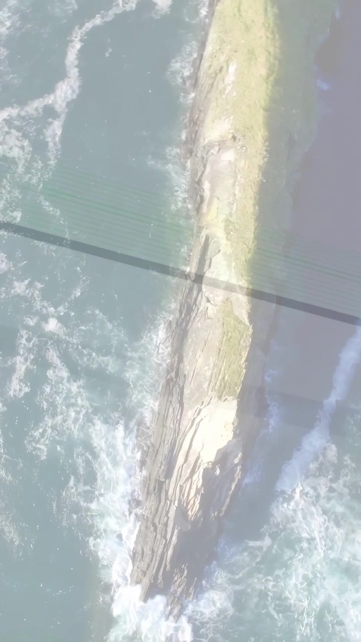 Video verticale: Telecamera che si inclina, onde che schiumano attorno a una lunga lingua di terra, l'ombra della griglia che si sposta, mostrando il flusso.