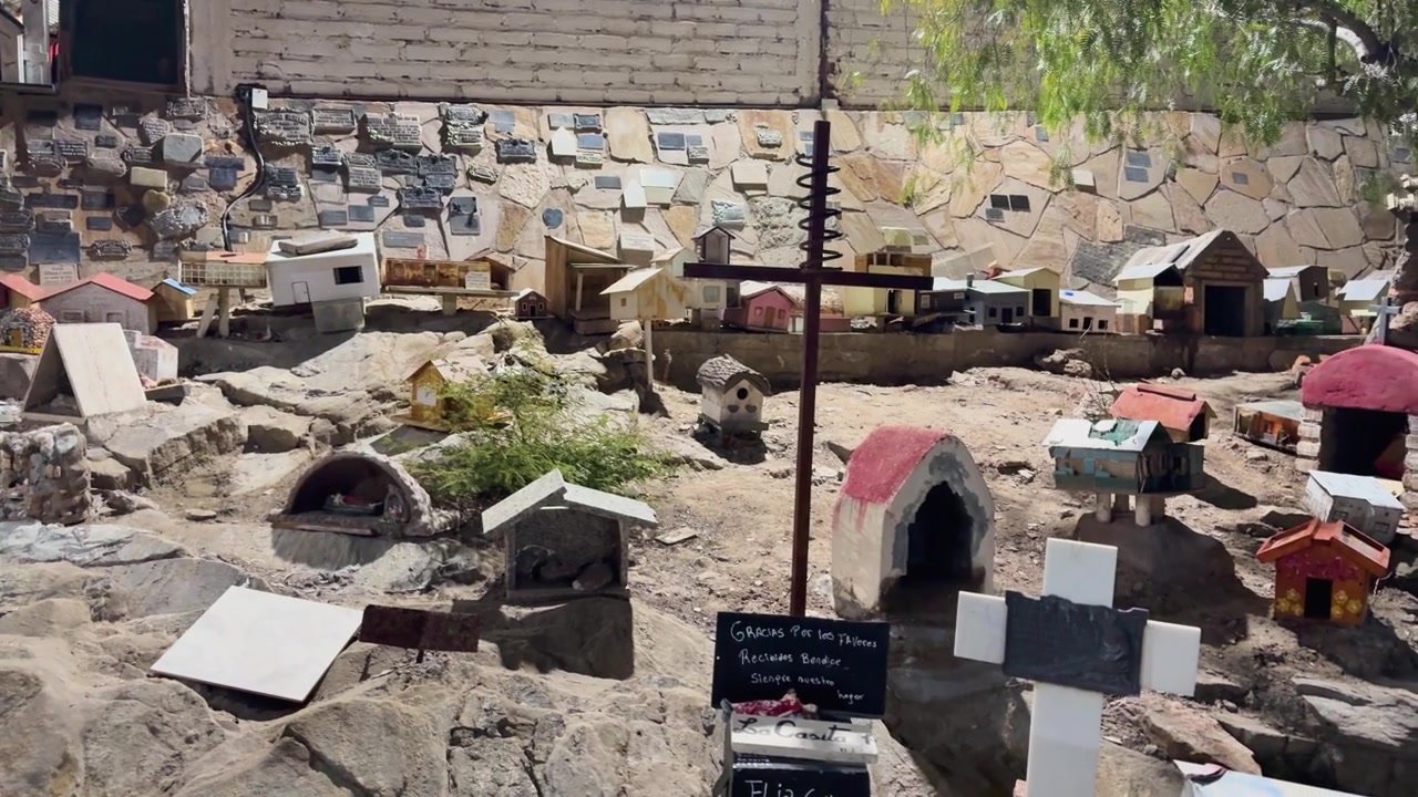 Panorámica diurna a mano alzada del santuario de la Difunta Correa en San Juan, Argentina, mostrando casas en miniatura, placas, cruces y ofrendas rústicas, iluminado por el sol y rodeado de rocas secas y monumentos conmemorativos.