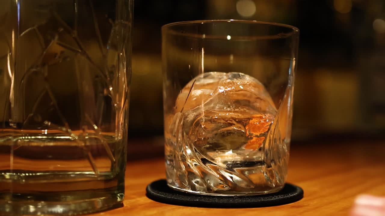Una mano fa scorrere un bicchiere di cristallo con una grande sfera di ghiaccio su un bancone da bar in legno.