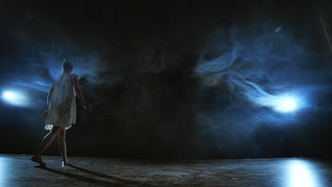 baila moderna mujer en un vestido blanco baila un ballet moderno salta en el escenario con humo en los reflectores azules.