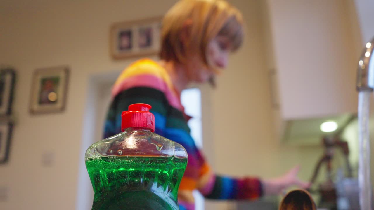 Soap bottle in focus, woman with brown hair cleans kitchen sink behind in blur