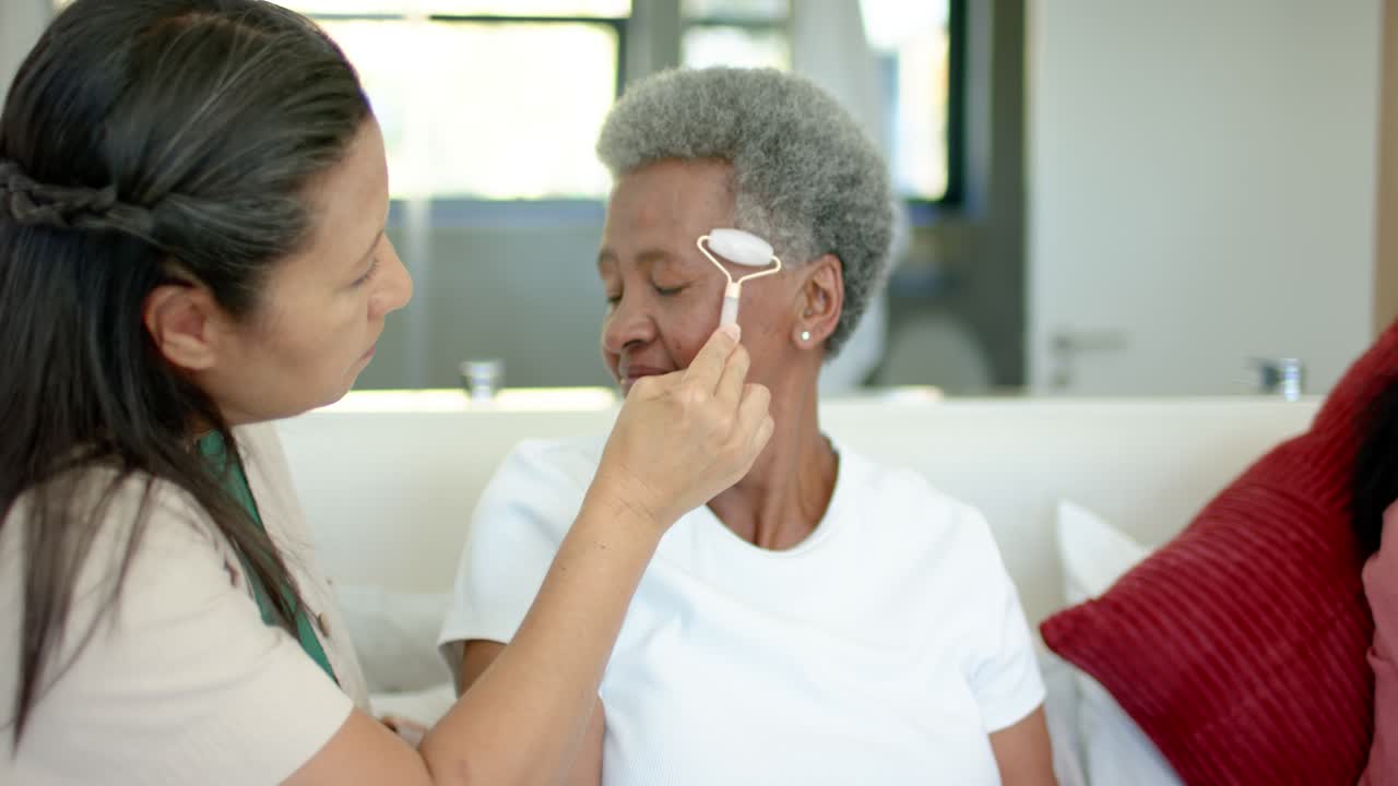 Cuidadora passando rolo facial rosa em mulher idosa após ela virar a cabeça, criando calma no sofá.