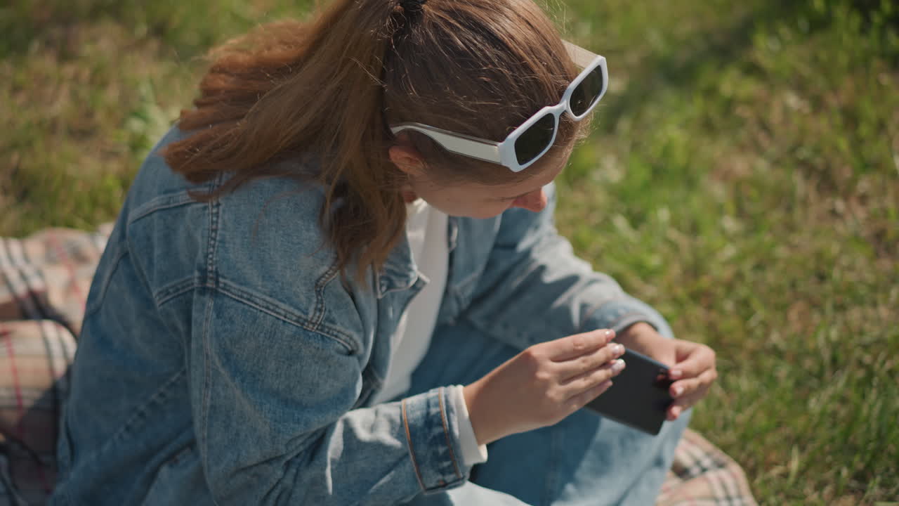 Kaukasische Frau untersucht im Freien ein kleines Gerät, sitzt auf einer Decke im Gras, Jeansjacke, weiße Sonnenbrille, praktisches Testen, Fokus auf Komponenten und Auspacken, Tutorial-Atmosphäre bei natürlichem Licht.