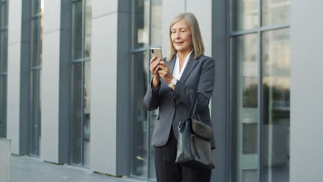 Mature-Businesswoman-In-Glases-Texting-And-Chatting-With-Smartphone-In-The-Street