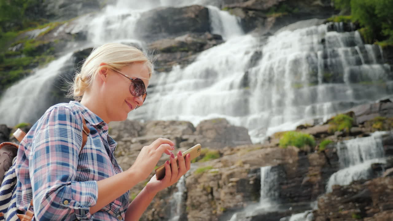 un viajero usa un teléfono inteligente contra el fondo de una cascada tvindefossen en noruega 4k video