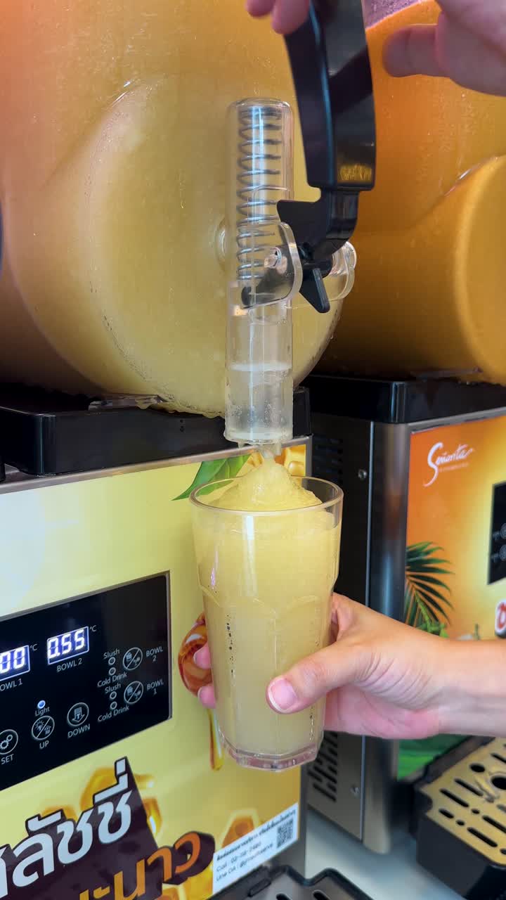Una toma de primer plano muestra una mano llenando un vaso con granizado de naranja de un dispensador.