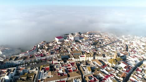 Slow-aerial-dolly-shot-to-the-back-over-the-old-town-medina-sidonia-in-spain-with-view-of-the-whole-old-town-with-white-buildings-with-occasional-red-roofs-and-the-clouds-in-the-background