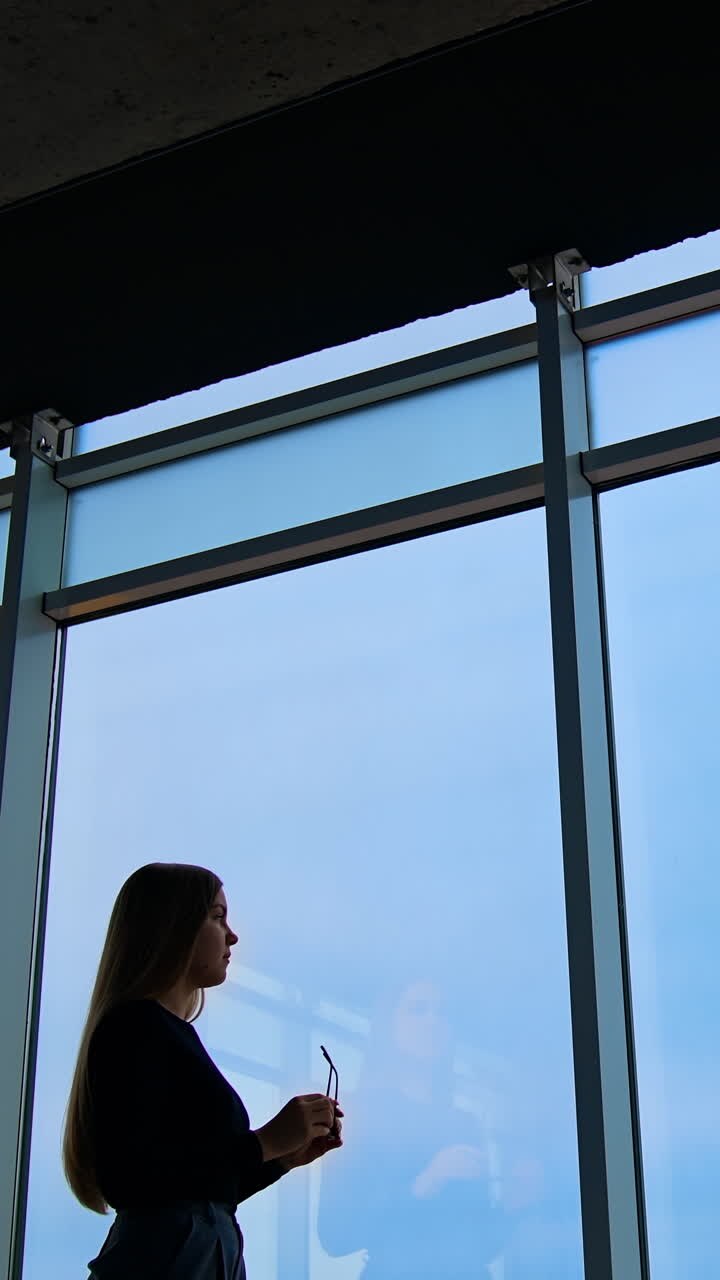 Business woman indoors. Tired female business worker in office putting off her glasses and looking through the large windows on blue sky background. Panoramic windows view. Vertical video