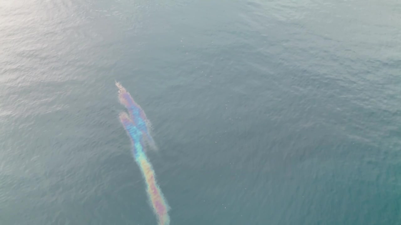 High-angle aerial drone clip showing a rainbow-colored iridescent oil slick and fuel spill contamination floating on the surface of calm ocean water, highlighting marine environmental pollution.
