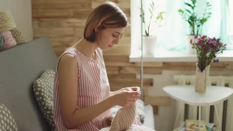 Craft-woman-sitting-on-couch-on-background-window-and-knitting-wool-scarf