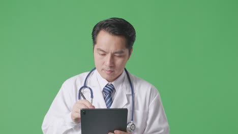 close up of asian male doctor with stethoscope taking note on a tablet while walking on green screen background in the hospital