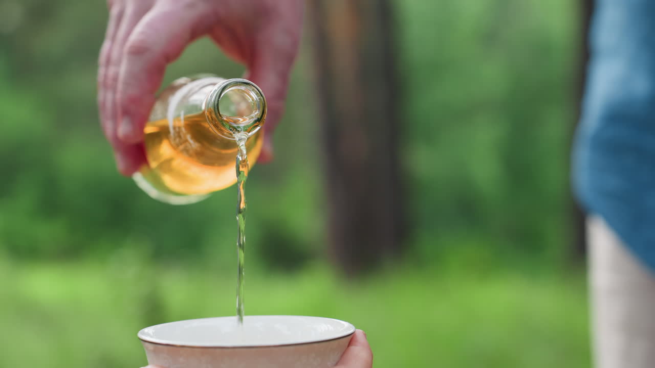 밝은 녹색 숲 속 야외에서 남성의 손이 유리병에서 흰색 컵으로 레모네이드를 부드럽게 따르는 모습이에요. 자연광 아래에서 상쾌한 여름 음료와 평온한 가족 피크닉 분위기를 보여줍니다.