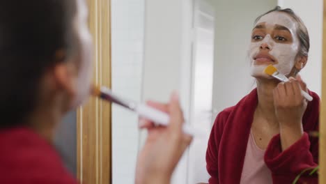 video de una feliz mujer biracial mirándose en el espejo aplicando una máscara facial con un cepillo en el baño