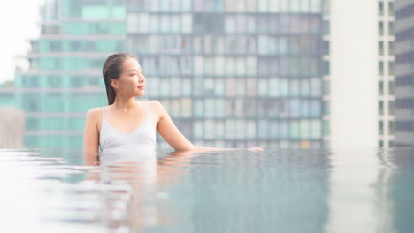 young asian woman relaxing on the edge of the rooftop infinity swimming pool overlooking the bangkok cityscape with tall skyscrapers at sunset