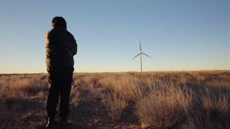 person stands in dry golden grass, facing a distant wind turbine on the patagonian plains while traffic quietly passes in the background under a clear evening sky, static camera