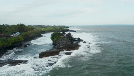 Toma-Aérea-Amplia-Del-Carro-Del-Templo-Vacío-Aislado-De-Tanah-Lot-Durante-Un-Clima-Peligroso.-Fuertes-Olas-Rompiendo-En-Un-Acantilado-Rocoso-Oscuro-Con-El-Famoso-Templo-Hindú-En-Bali,-Indonesia