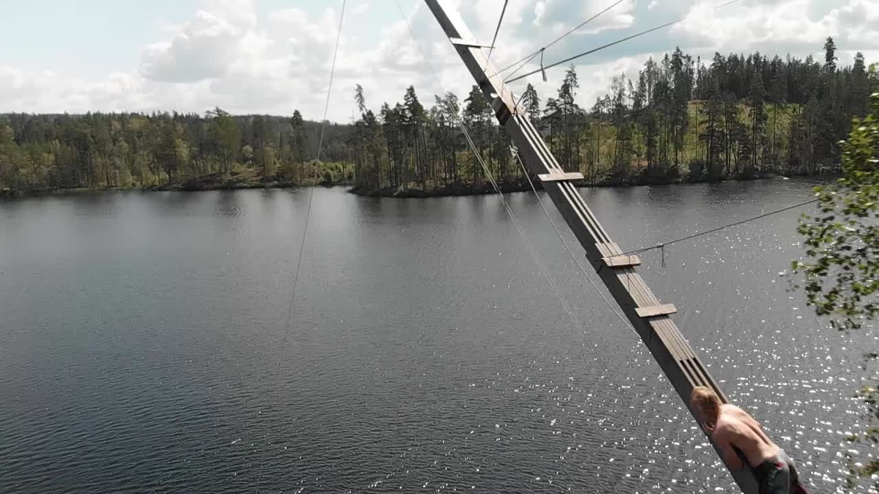 Adult Male Jumping Off Wooden Platform On Rope Swing Into Lake. Follow Shot