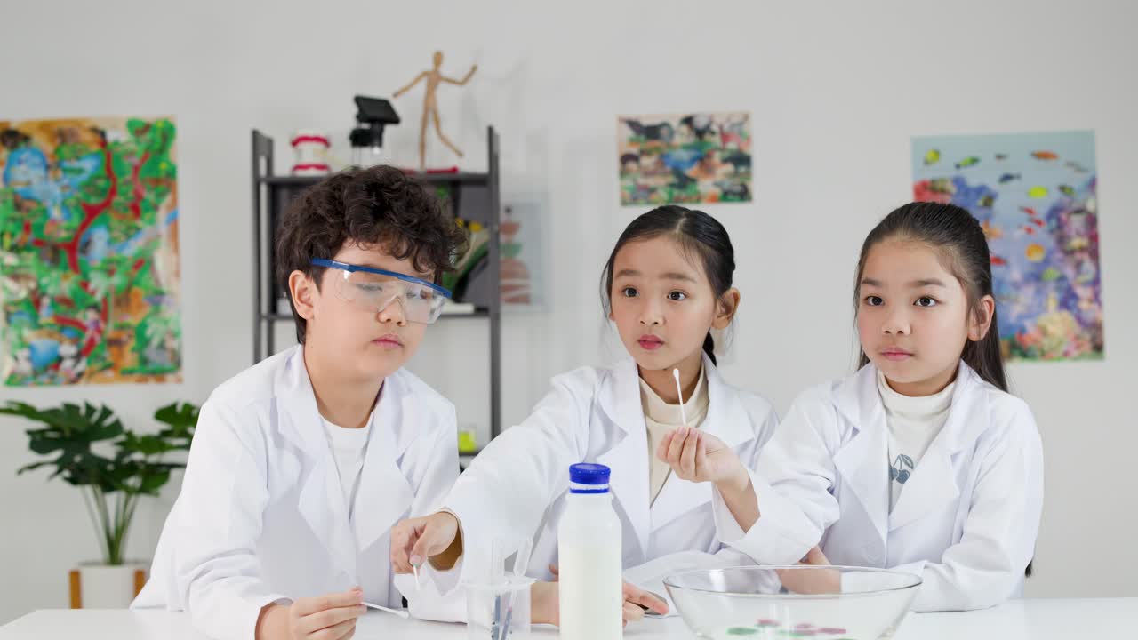 白衣を着た3人のアジア人の子供たちが、牛乳と食用色素を使った化学実験を行っている