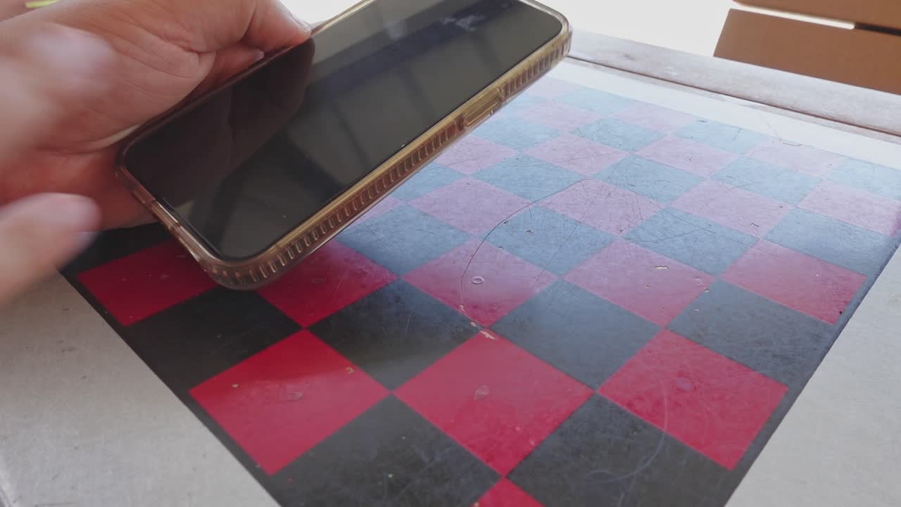 A person picks up a smartphone from a park table with a black and red checkerboard pattern