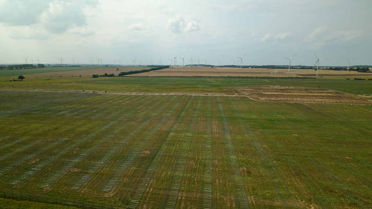 Ladang Hijau Luas untuk Ladang Panel Surya dengan Turbin Angin di Latar Belakang Dekat Zwartowo di Polandia.