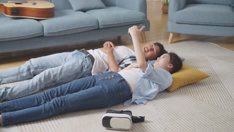 asian teen couple having video call on smartphone while lying on carpet on the floor at home. waving hands, speaking, and laughing