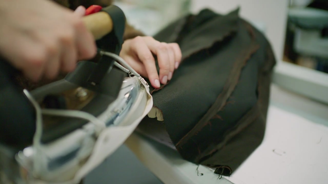 Nahaufnahme eines Schneiders, der ein professionelles Dampfbügeleisen auf schwarzem Stoff in einem Modeatelier verwendet.