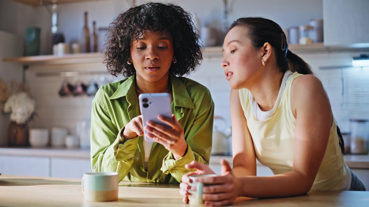 Multiethnische Freunde teilen sich ein Telefon in der Küche. Zwei Frauen surfen mit dem Smartphone bei Kaffee