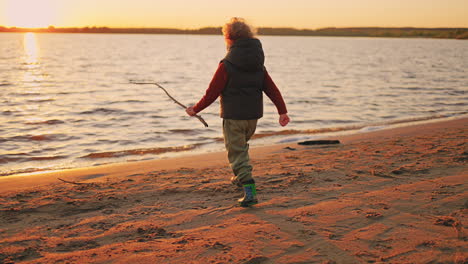 Fröhlicher-Kleiner-Lockiger-Junge-Rennt-Bei-Sonnenuntergang-Zum-Wasser-Des-Flusses-Und-Spaziert-Am-Herbst--Oder-Frühlingsabend-An-Der-Sandküste