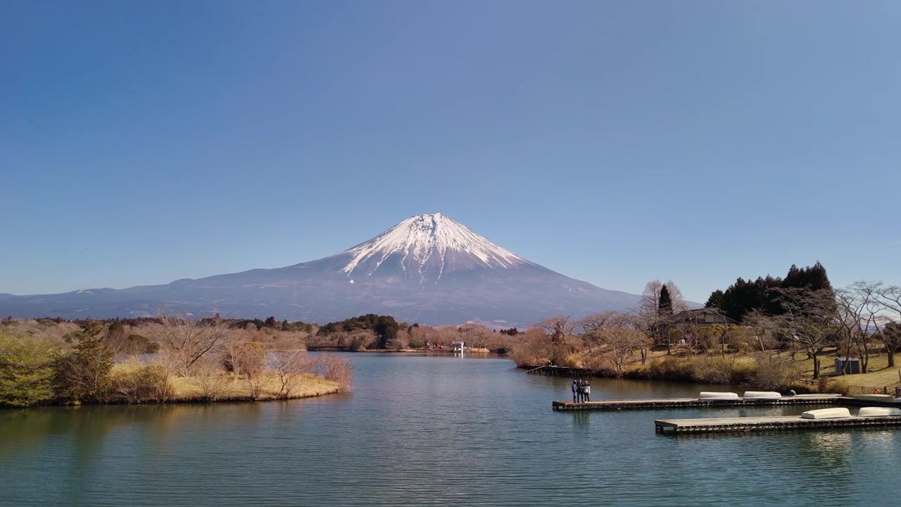 Pittoresca veduta grandangolare del Monte Fuji che si erge su un lago calmo, con turisti che ammirano il paesaggio da un molo.