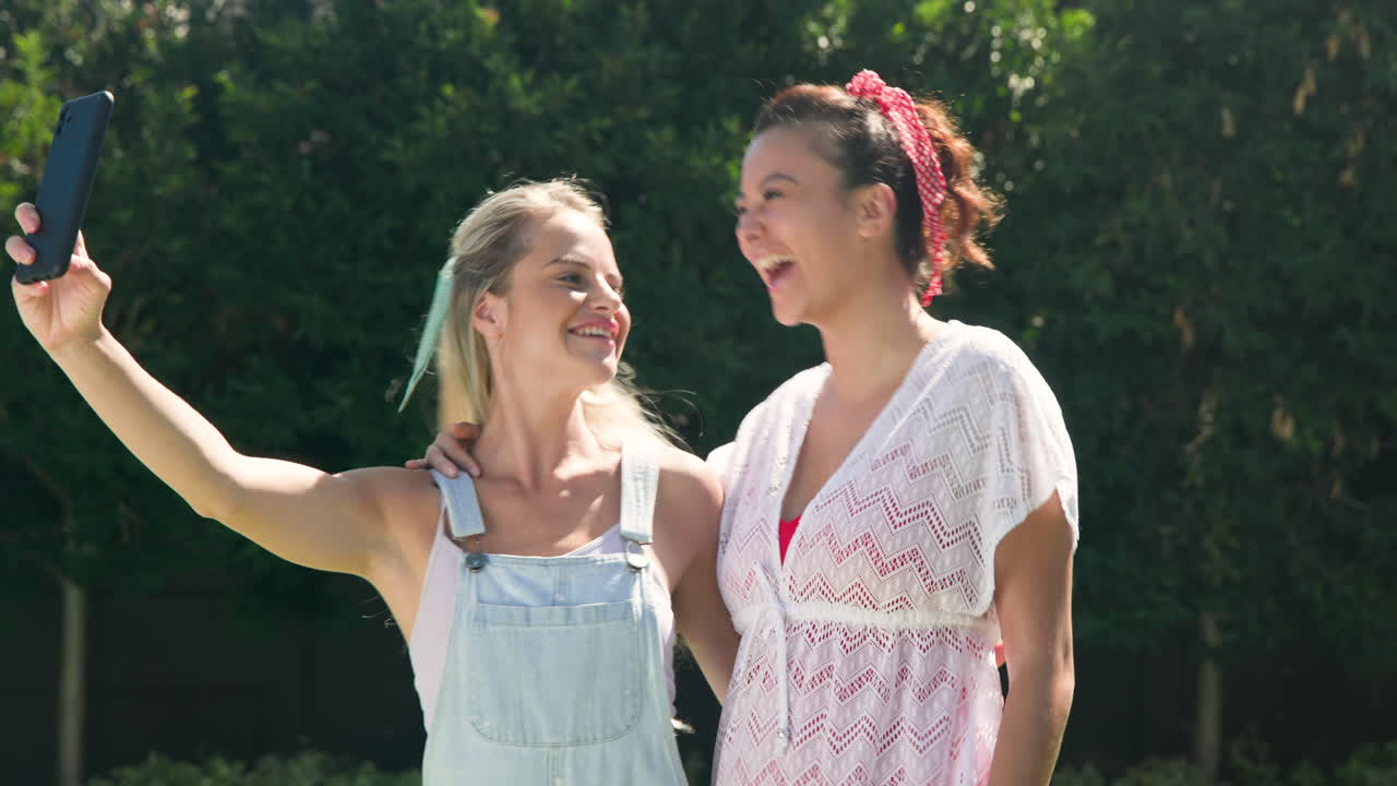 Two female friends enjoying sunny day by pool, looking at smartphone together