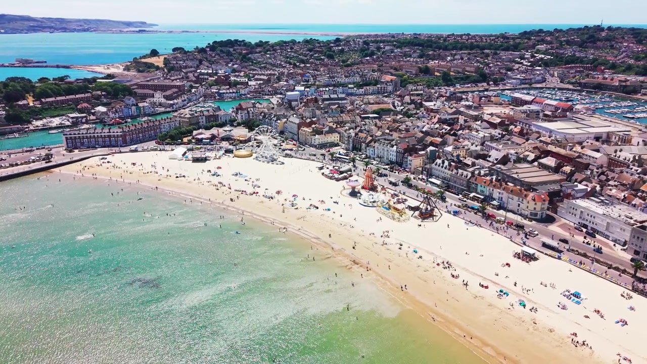 Ripresa con drone della spiaggia di Weymouth con giostre del luna park, folle di vacanzieri e il lungomare