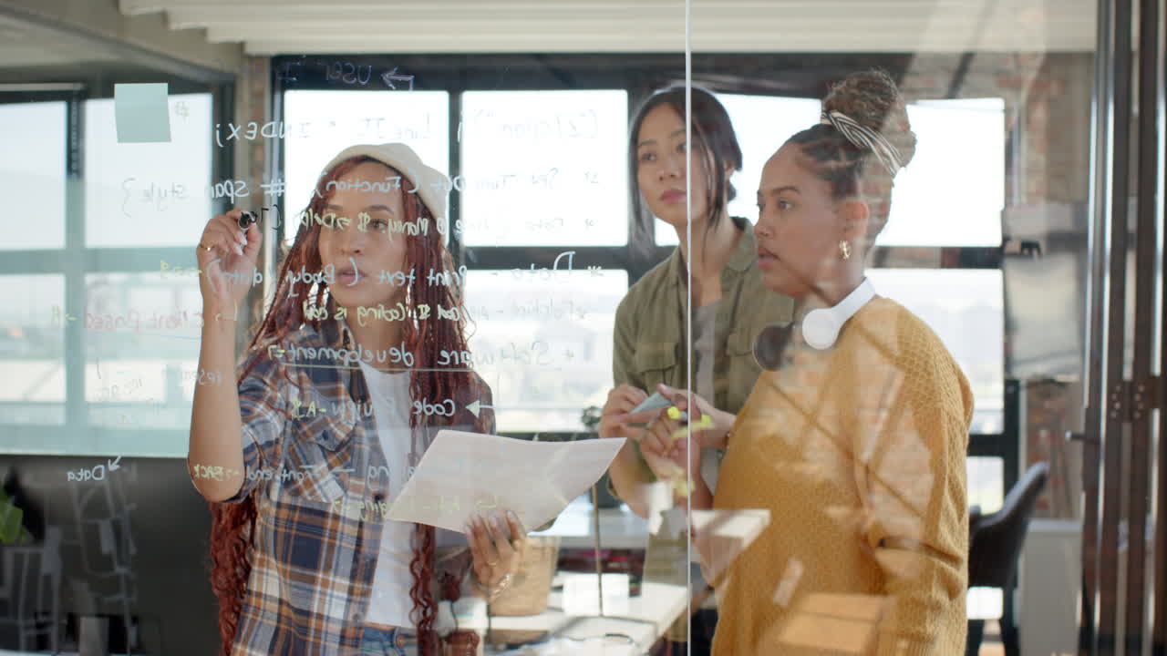 Diverse female team collaborating on coding project, writing on glass board in modern office